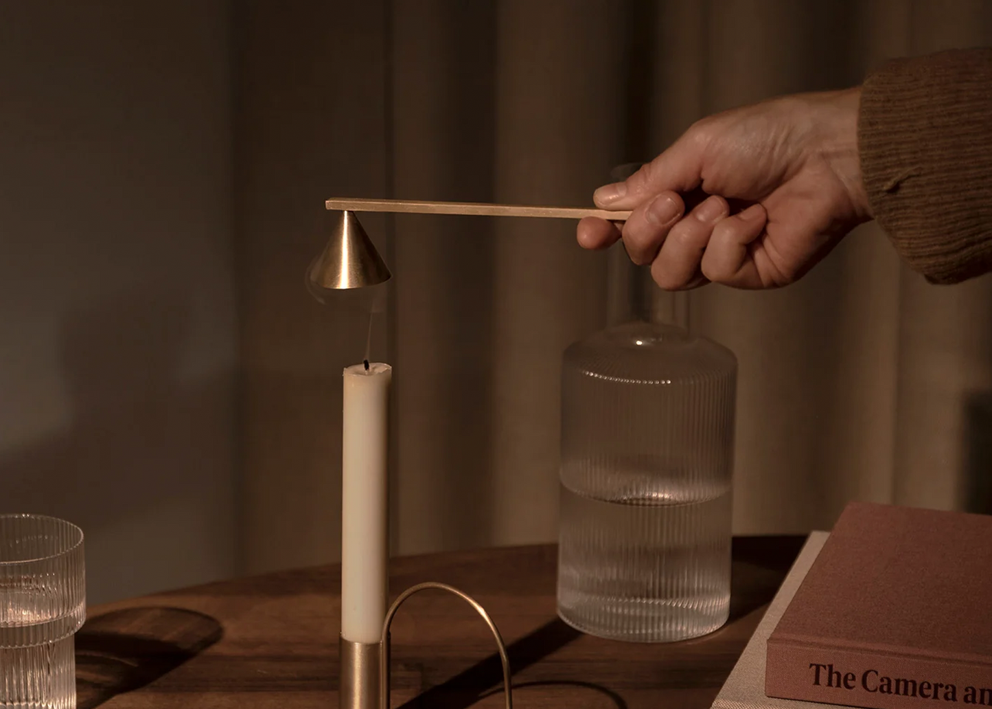 A brass candle extinguisher by Ferm Living with a cone extinguisher and long straight handle as sold by Woodland Mod being held by a hand while putting out a flame of a long candle