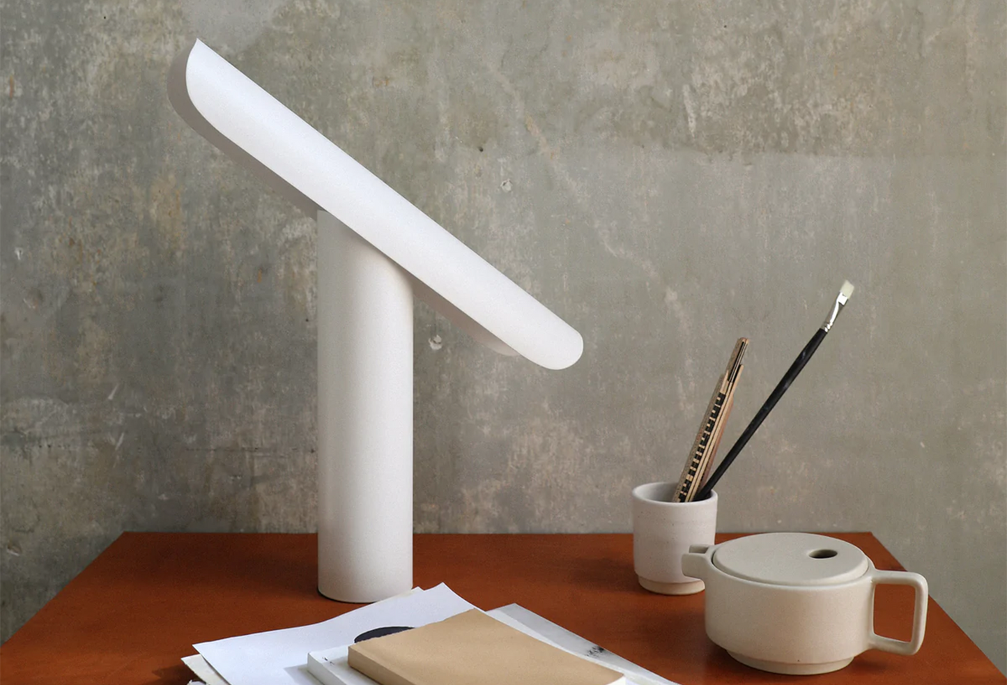 A white T-shaped FRAMA T-Lamp as sold by Woodland Mod on a desk with papers, pens, and a tea pot.