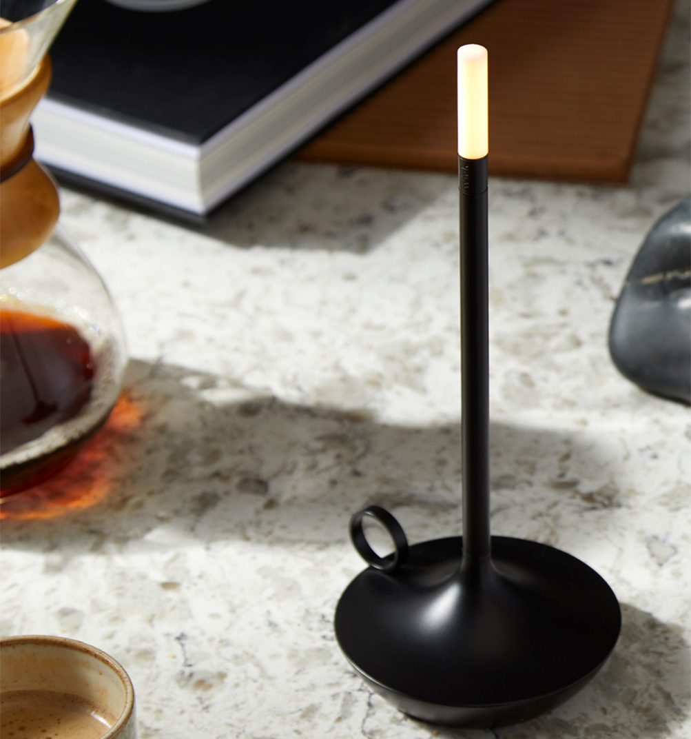 A black wick lamp by Graypants illuminated on a marble countertop next to desk supplies