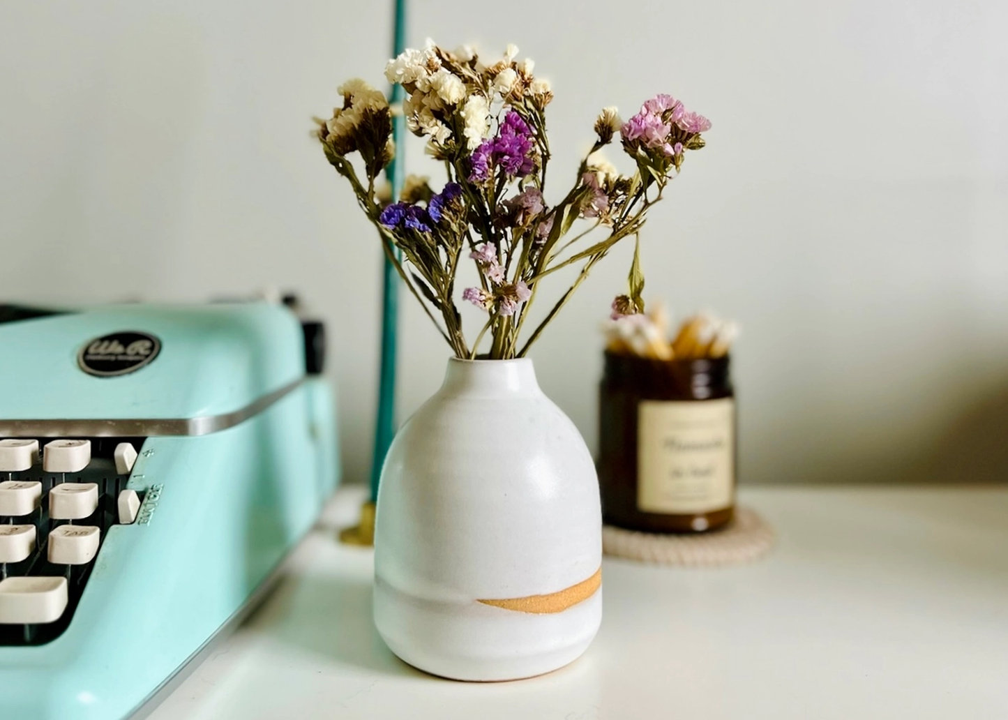 
                  
                    A white ceramic vase with a small orange accent band, holding dried flowers, styled on a tabletop next to a mint green typewriter and a small amber jar.
                  
                