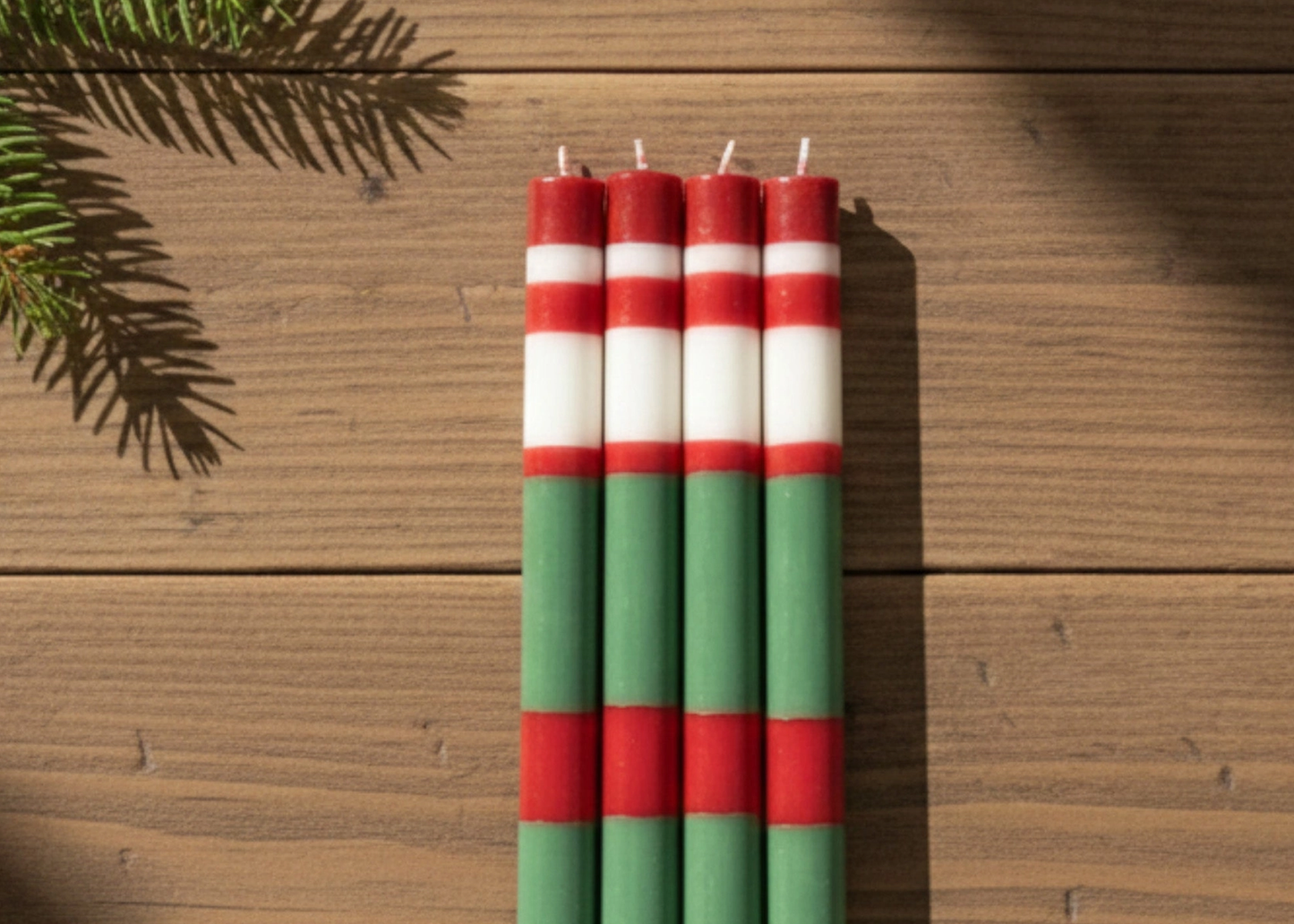 
                  
                    Four striped taper candles with red, white, and green horizontal bands, standing upright on a rustic wooden surface with pine branches in the corner.
                  
                