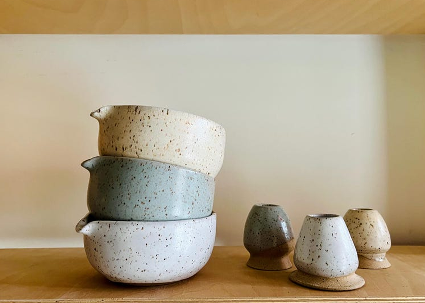 
                  
                    A set of handmade ceramic tableware displayed on a wooden shelf against a neutral beige wall. The set includes a stack of three speckled bowls in cream, sage green, and off-white, alongside three matching small ceramic cups or vases.
                  
                