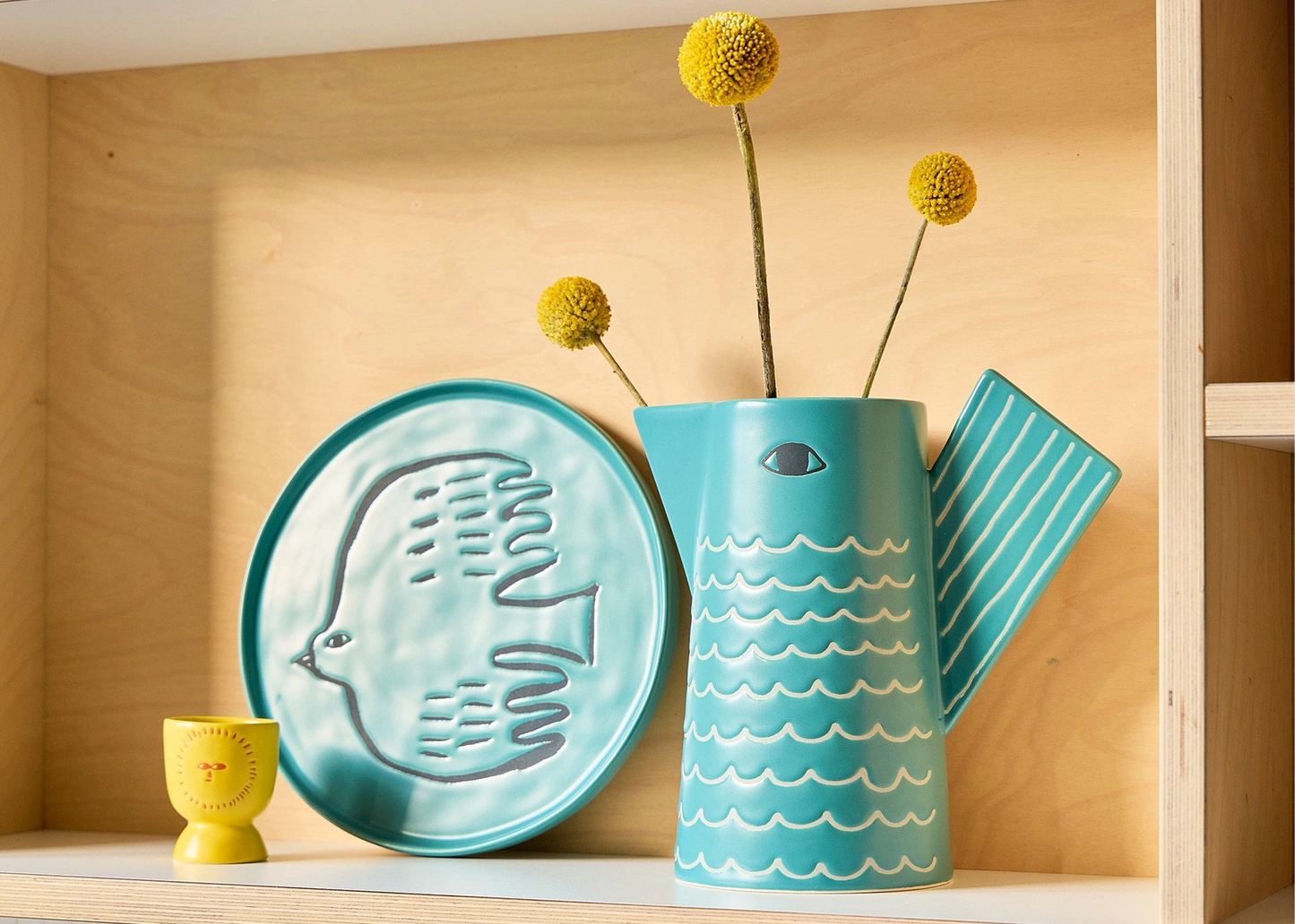 
                  
                    A turquoise ceramic pitcher vase with a handle, shown on a wooden shelf alongside a matching round plate and a small yellow cup. The vase has a wave pattern and holds three yellow pom-pom flowers.
                  
                