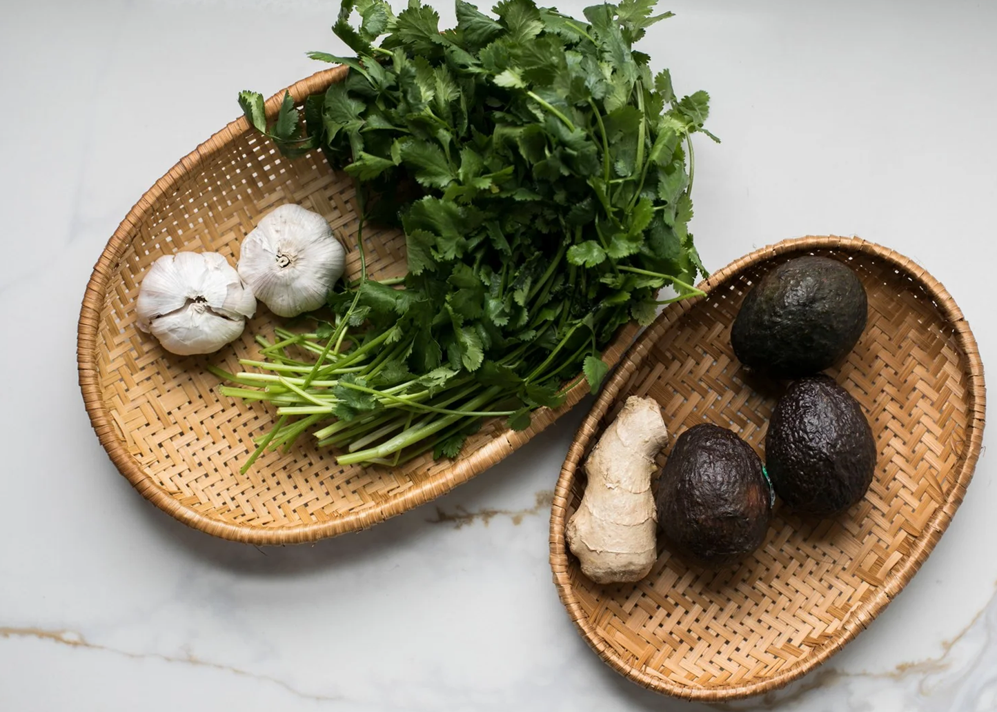 
                  
                    Fresh ingredients arranged on two woven oval trays on a light marble surface: a pile of green cilantro, two white garlic bulbs, a piece of ginger root, and three dark avocados.
                  
                