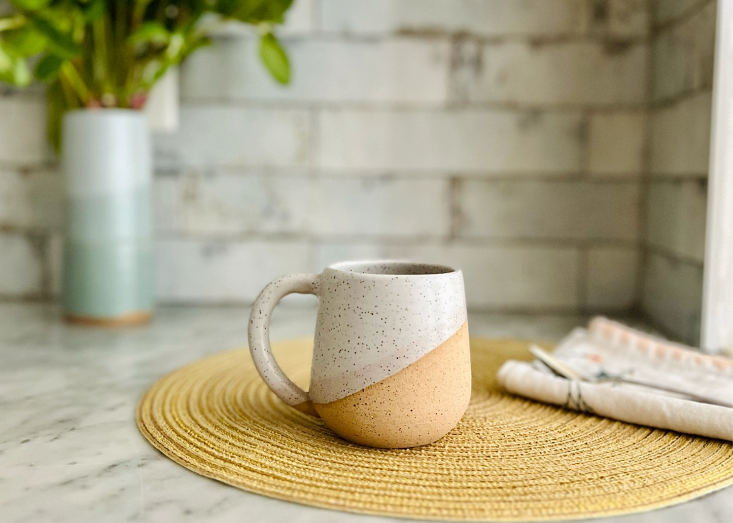 
                  
                    A ceramic mug with a diagonal two-tone design in speckled cream and warm peach-orange, resting on a round woven placemat on a marble countertop.
                  
                