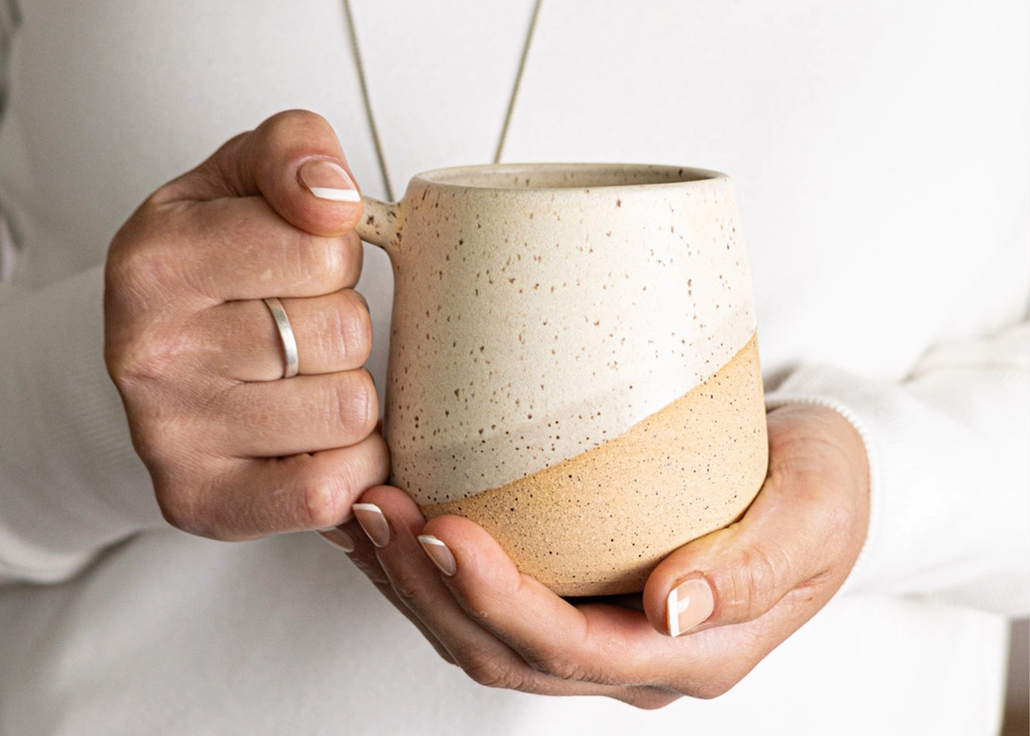 
                  
                    A person holding a ceramic mug with both hands. The mug has a speckled cream and light tan color-blocked design, with a cream upper half and a warm beige lower half.
                  
                