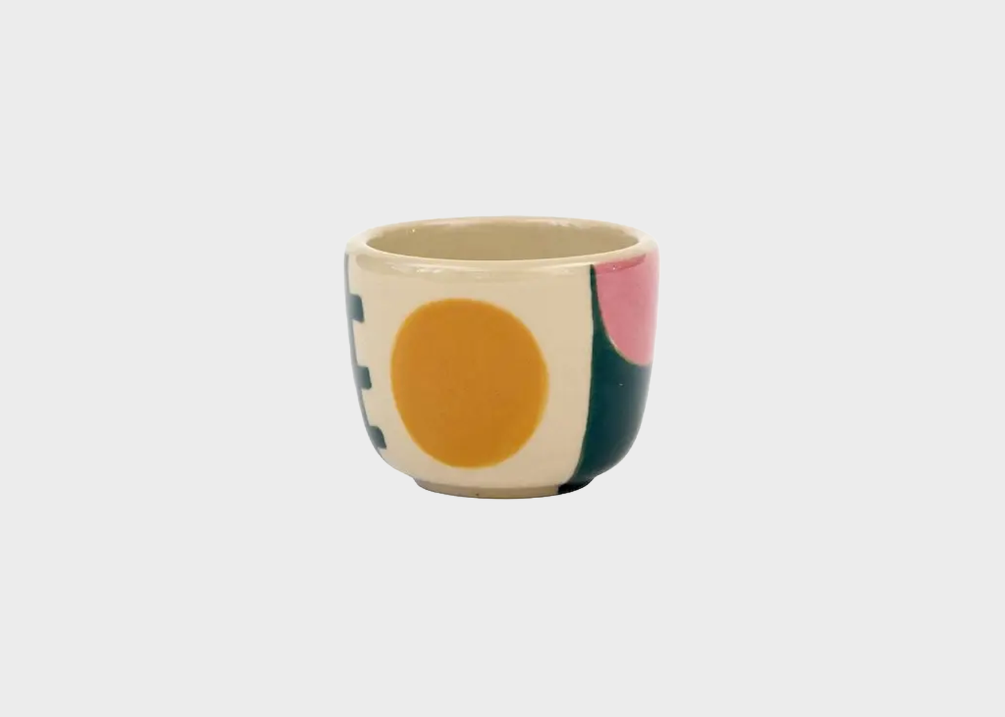 
                  
                    A small ceramic cup with a cream base and a colorful abstract geometric pattern featuring a large mustard yellow circle, a pink shape, a dark green shape, and a blue shape, set against a plain light gray background.
                  
                