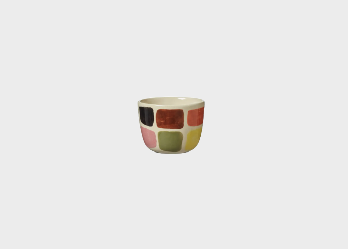 
                  
                    A small ceramic cup with a retro patchwork design of colored square tiles on a light beige background, centered against a solid light gray backdrop.
                  
                