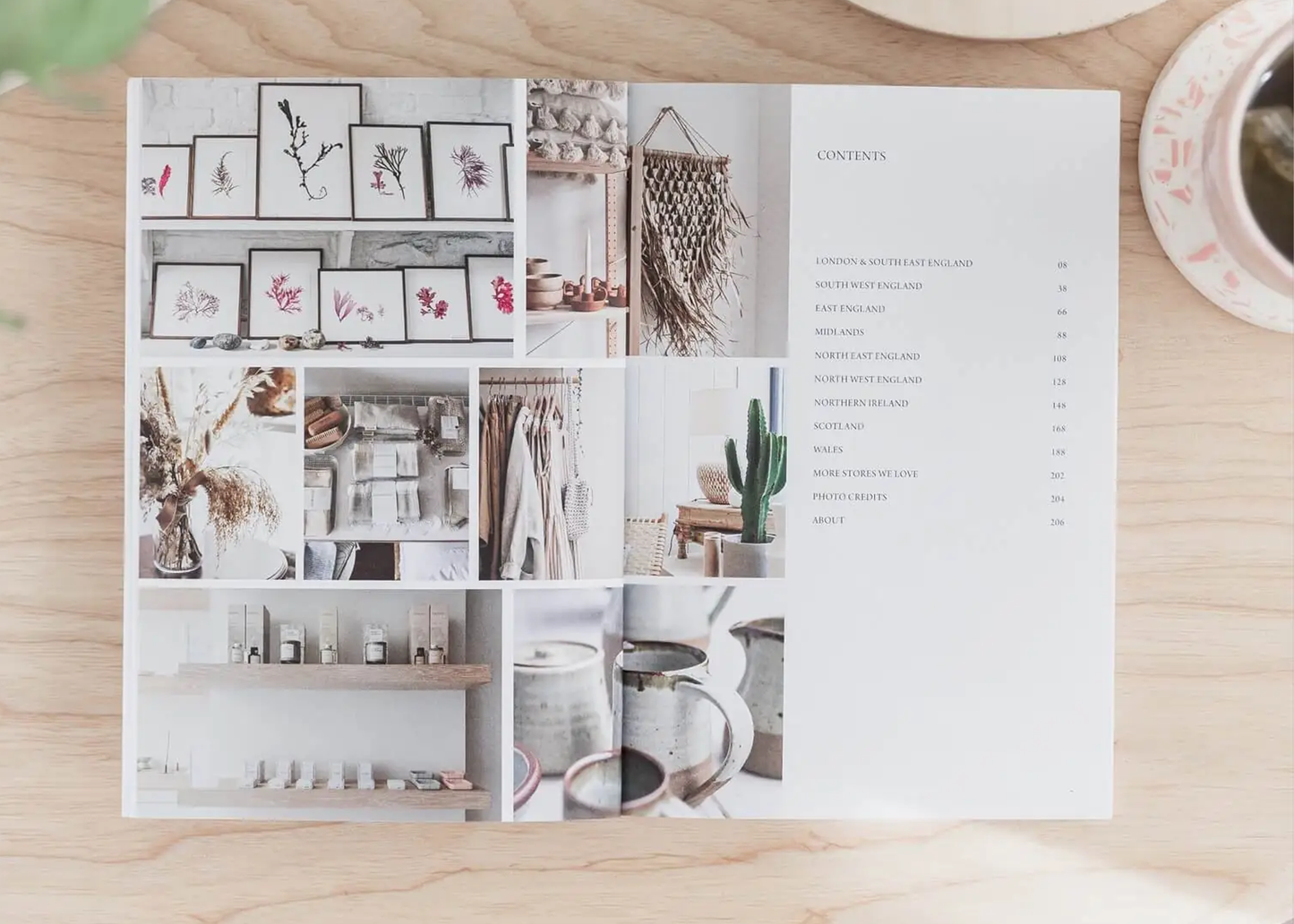 
                  
                    An open book spread laid flat on a light wood surface, showing a collage of interior design photos and a contents page. A cup and saucer are partially visible at the top right, and the catalog is the main product-like subject in this styled flat lay.
                  
                