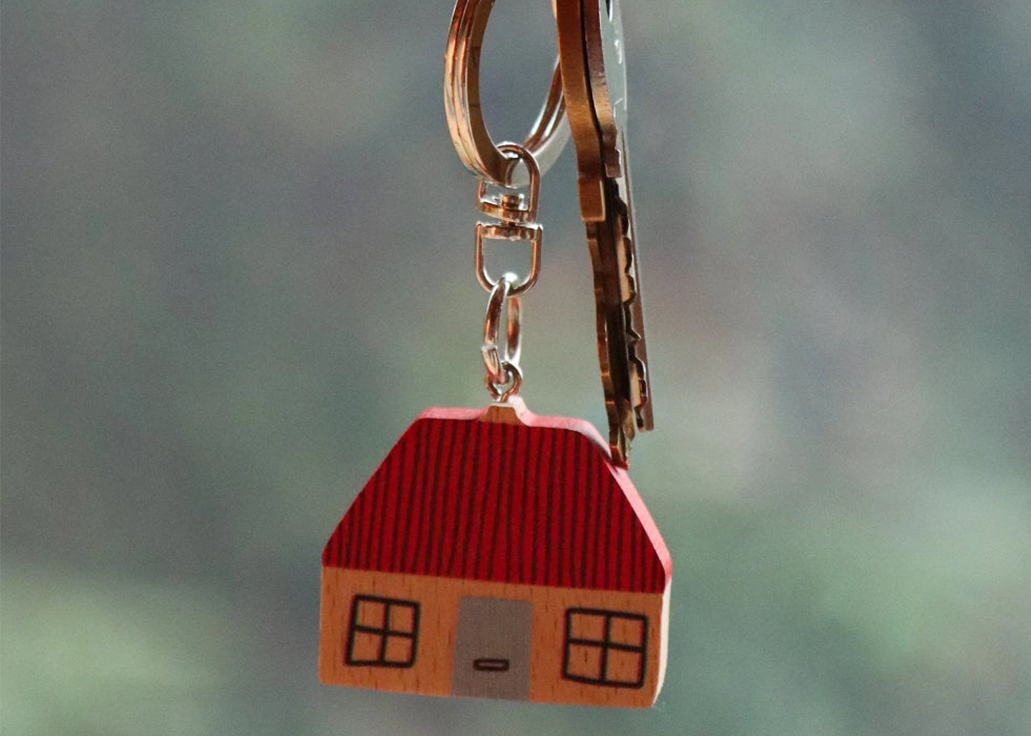 
                  
                    A small house-shaped keychain charm with a red roof and tan body, attached to a metal keyring with keys, shown against a blurred green-gray background.
                  
                