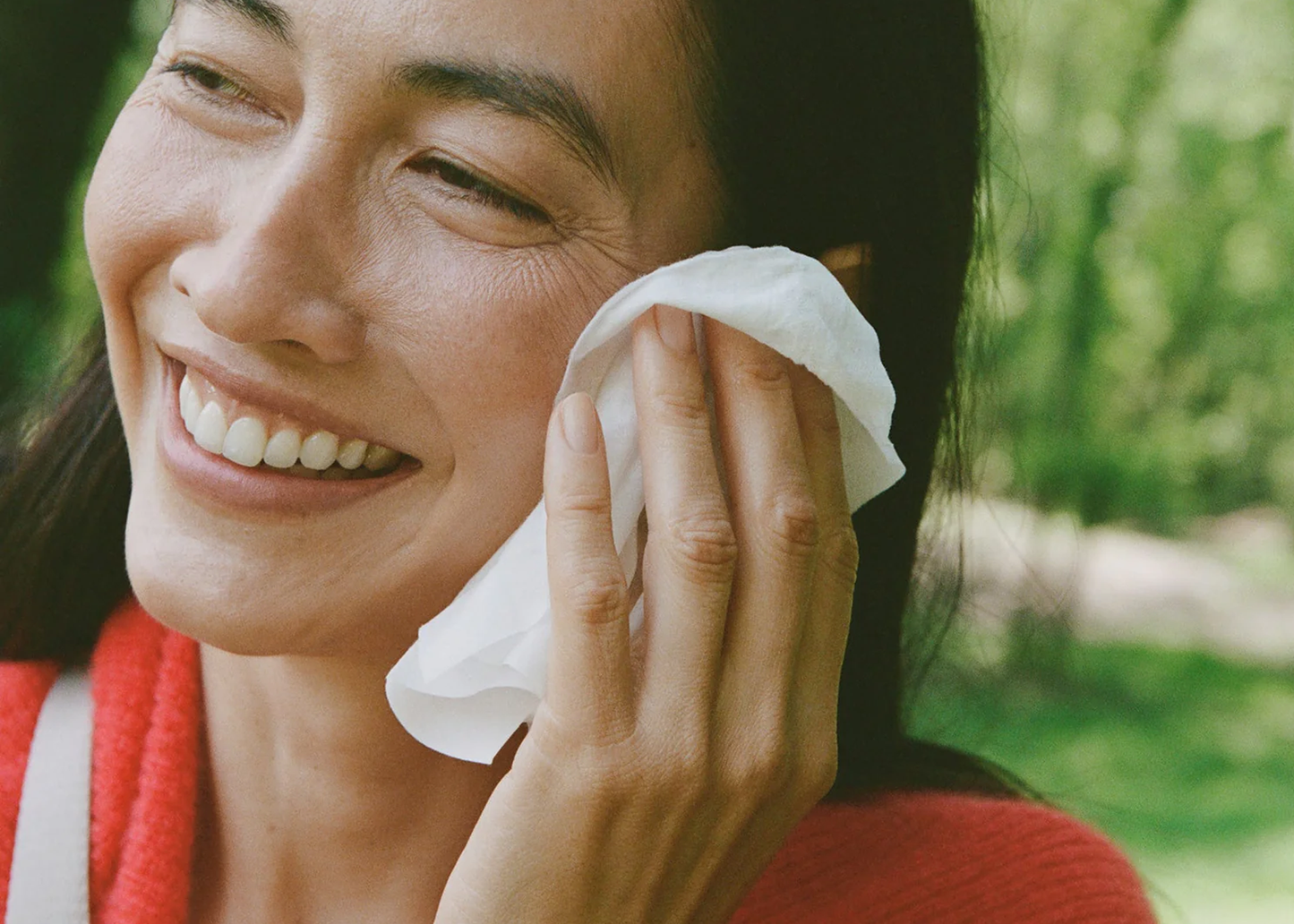 
                  
                    A close-up lifestyle shot of a woman outdoors, smiling and wiping her face with a white tissue or cotton pad. She is wearing a red knit top, with a soft-focus green garden background.
                  
                