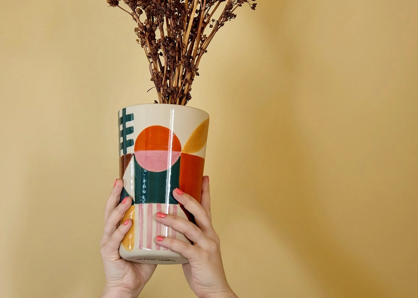 
                  
                    A person's hands holding a ceramic planter with a colorful geometric pattern, containing dried brown branches, set against a solid warm beige background.
                  
                