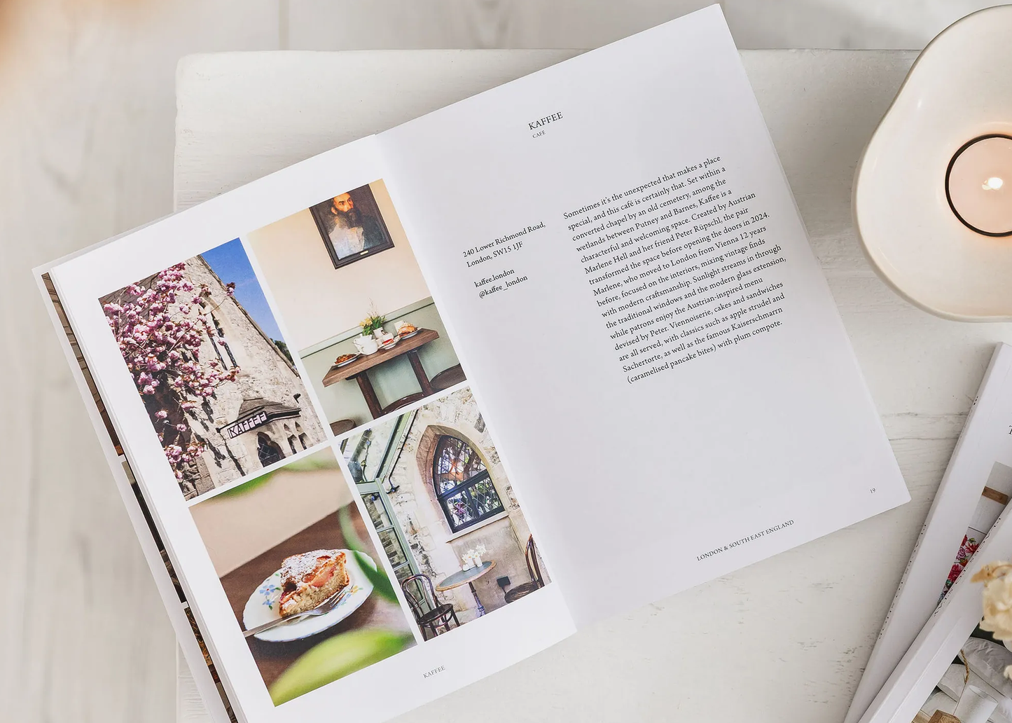
                  
                    An open book spread laid on a light marble surface, showing a page with text and several small architectural and interior photographs. A cream candle holder is partially visible on the right, and the scene is styled in a soft, clean flat-lay arrangement.
                  
                