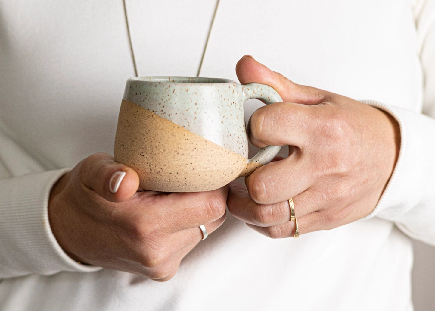 
                  
                    A close-up of a person holding a ceramic mug with a speckled light gray upper half and a warm beige lower half. The person is wearing a white long-sleeve top and rings on their fingers.
                  
                