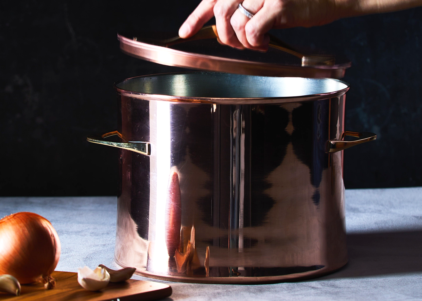 
                  
                    A large copper stockpot with a lid being lifted by a hand, set on a gray surface with an onion and garlic in the foreground against a dark background.
                  
                