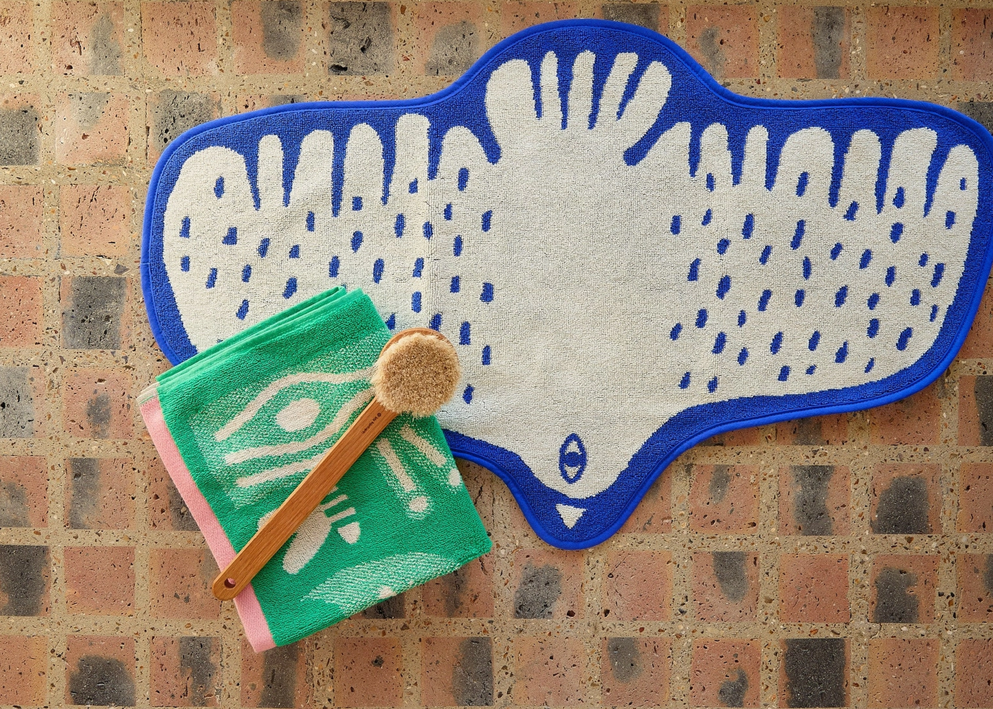 
                  
                    A white textured bath mat with a blue outline and blue dotted pattern, styled on a brick floor next to a folded green towel with a wooden scrubber.
                  
                
