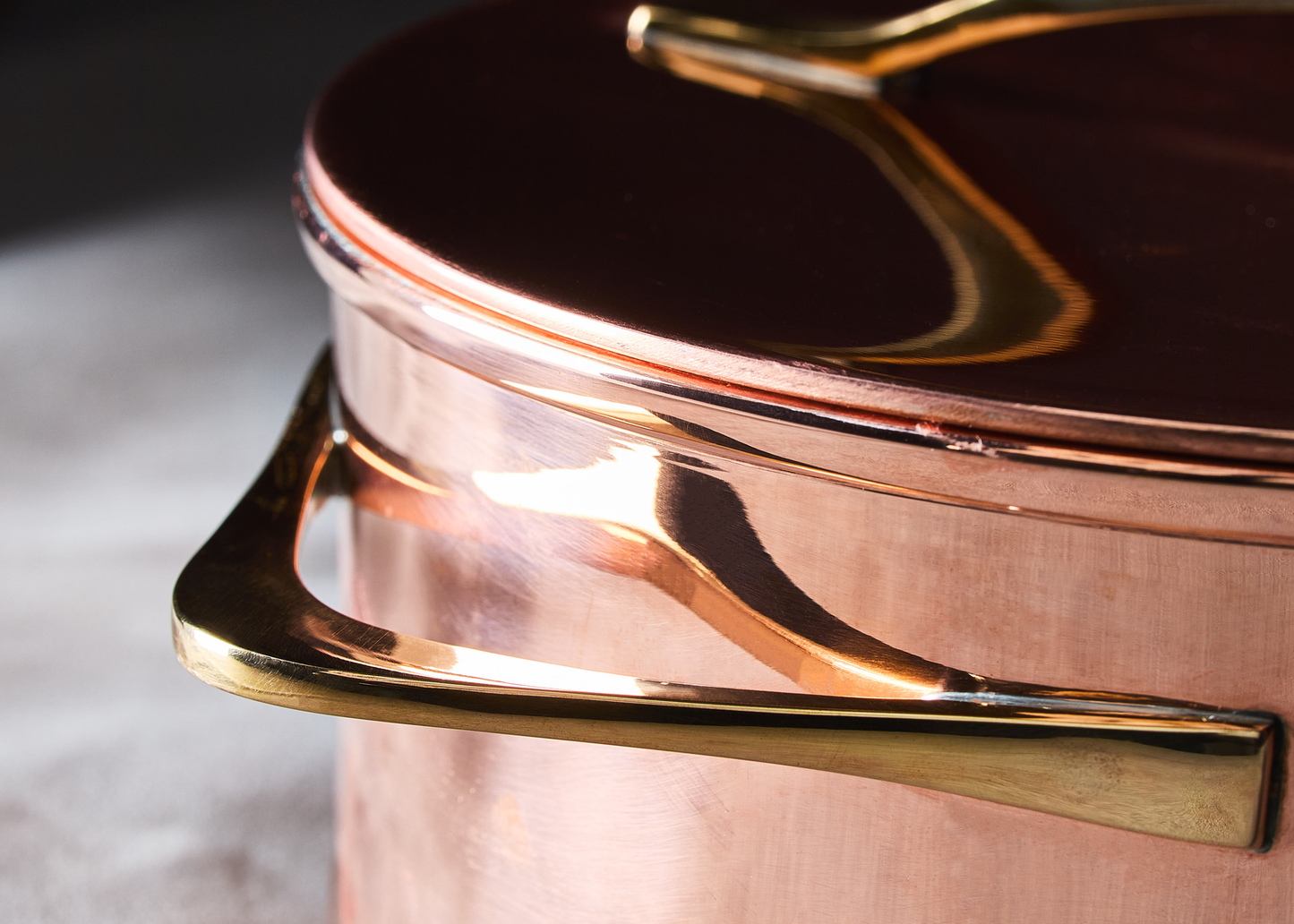 
                  
                    Close-up of a copper pot with a shiny gold-toned metal handle and lid, set against a dark and light gray blurred background.
                  
                
