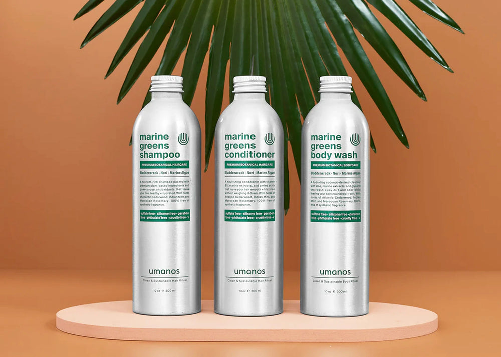 Umanos Marine Greens Sustainable Shower Set containing three aluminum bottles with eco friendly shampoo, conditioner, and body wash set in front of an orange background with a palm frond in the back.