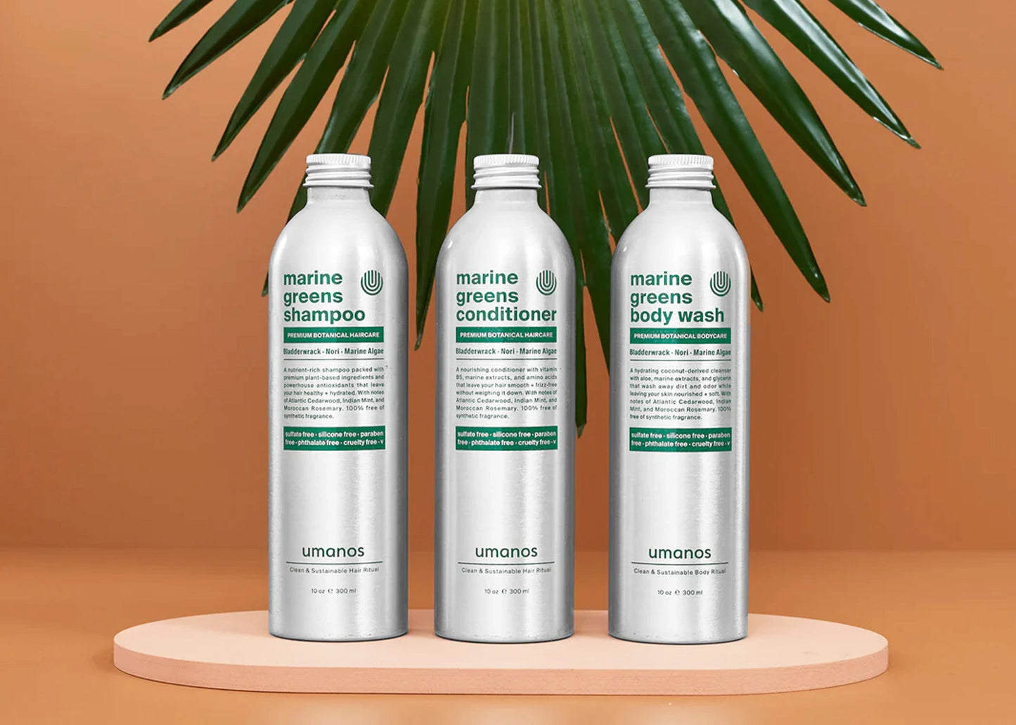 
                  
                    Umanos Marine Greens Sustainable Shower Set containing three aluminum bottles with eco friendly shampoo, conditioner, and body wash set in front of an orange background with a palm frond in the back.
                  
                