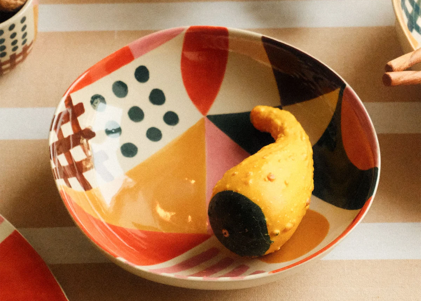 
                  
                    Decorative ceramic bowl with geometric pattern containing a yellow fruit on a striped surface.
                  
                