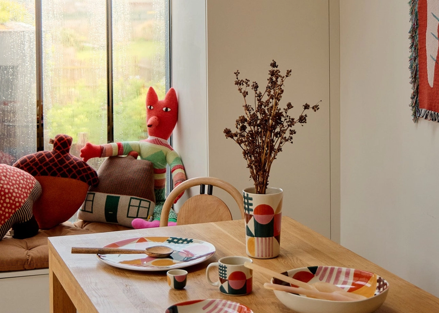 
                  
                    A styled dining corner with a light wood table set with matching ceramic dishes, cups, and a vase holding dried stems, alongside colorful pillows and a red plush fox toy near a window.
                  
                