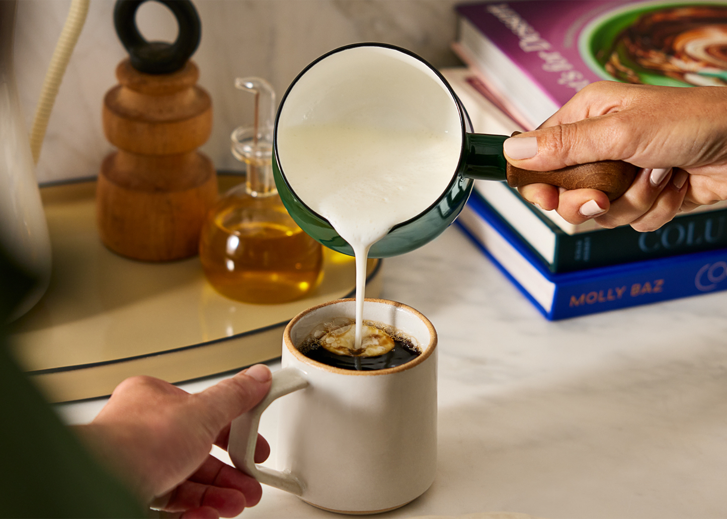 
                  
                    Person pouring milk into a coffee mug with a blurred background of books and kitchen items.
                  
                