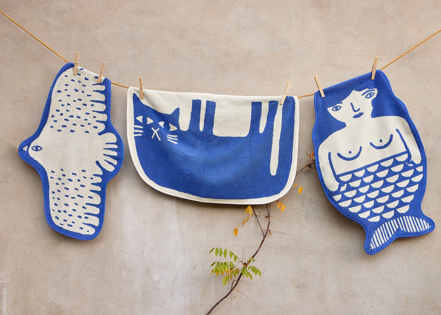 
                  
                    Three blue and white fabric hanging cloths with hand-drawn style illustrations of a bird, a cat, and a mermaid, suspended by a string against a beige wall.
                  
                
