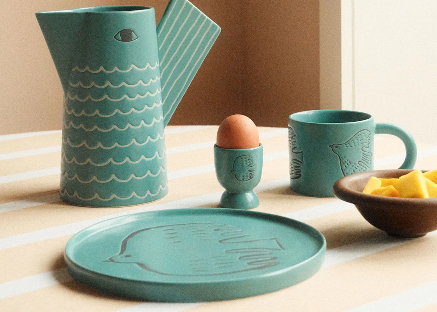 Teal ceramic kitchenware set including pitcher, mug, plate, and egg cup on a table.