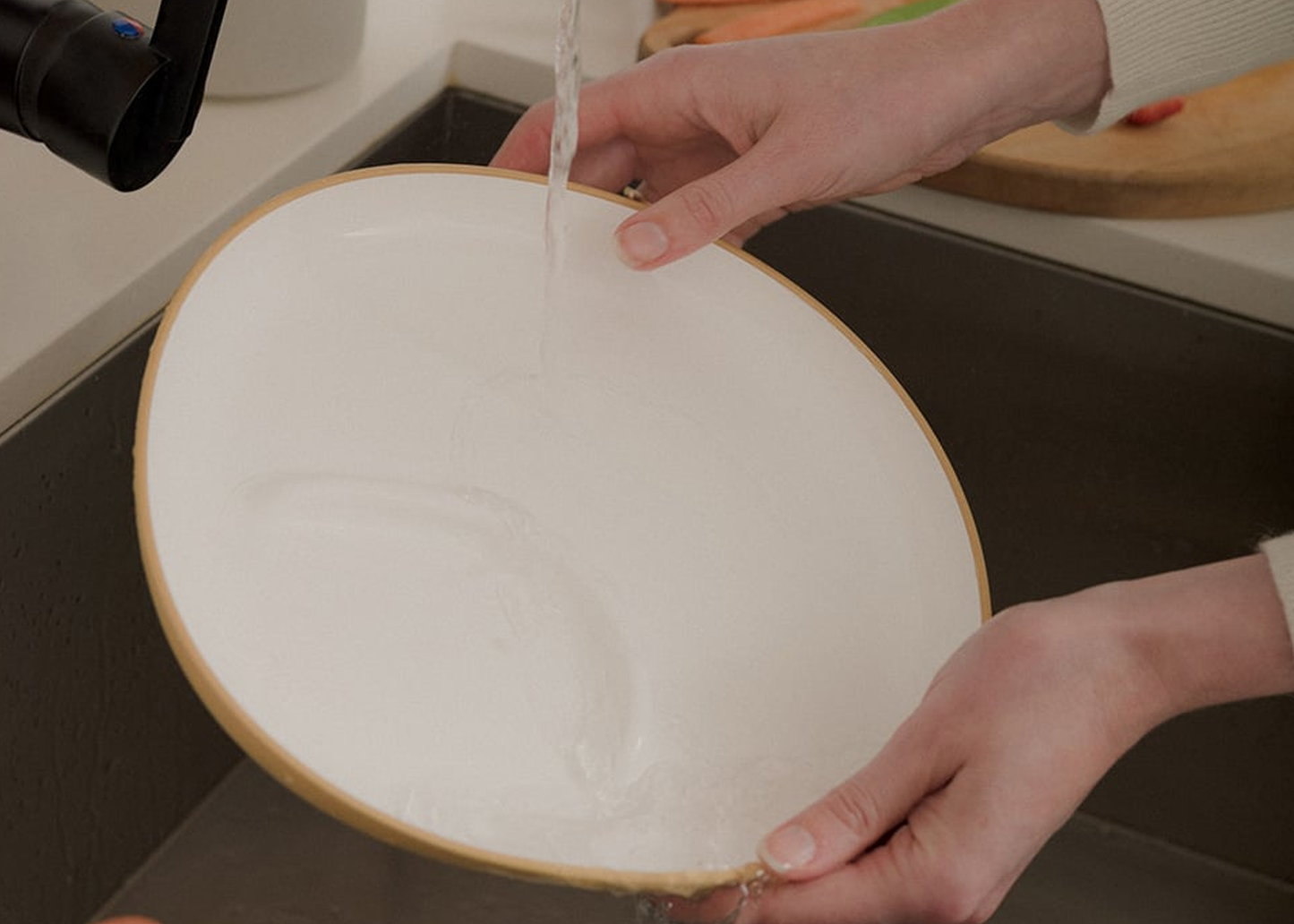 
                  
                    Person washing a white plate with gold rim under running water in a kitchen sink.
                  
                
