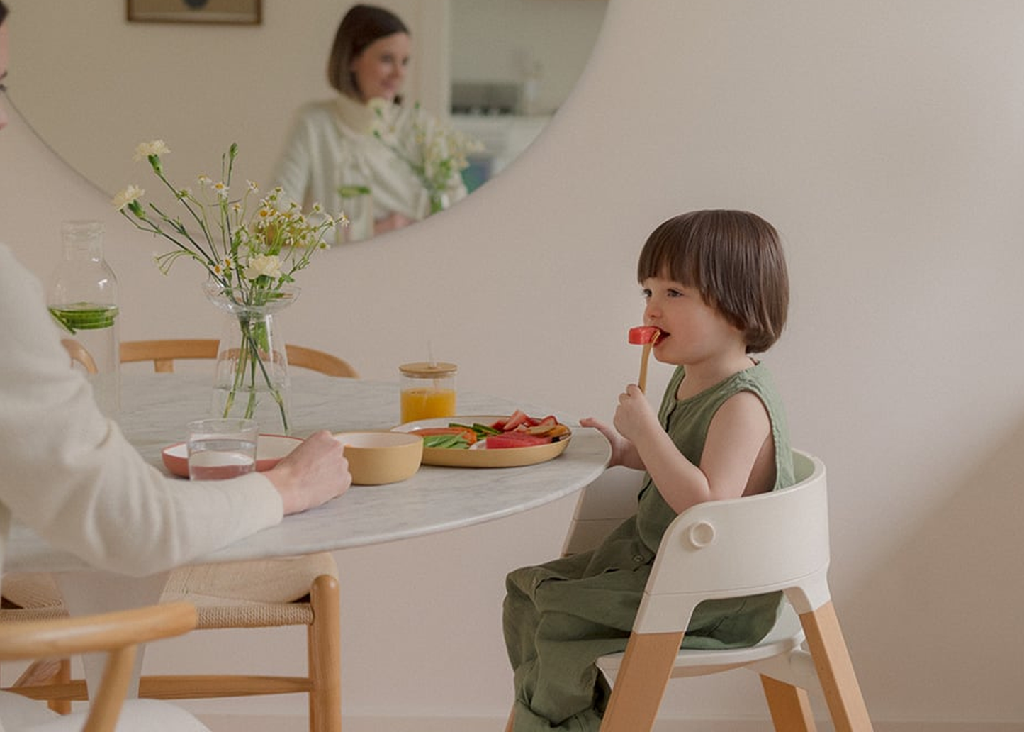 
                  
                    Child sitting at a dining table with a plate of food and a glass of juice, with a blurred background of a family.
                  
                