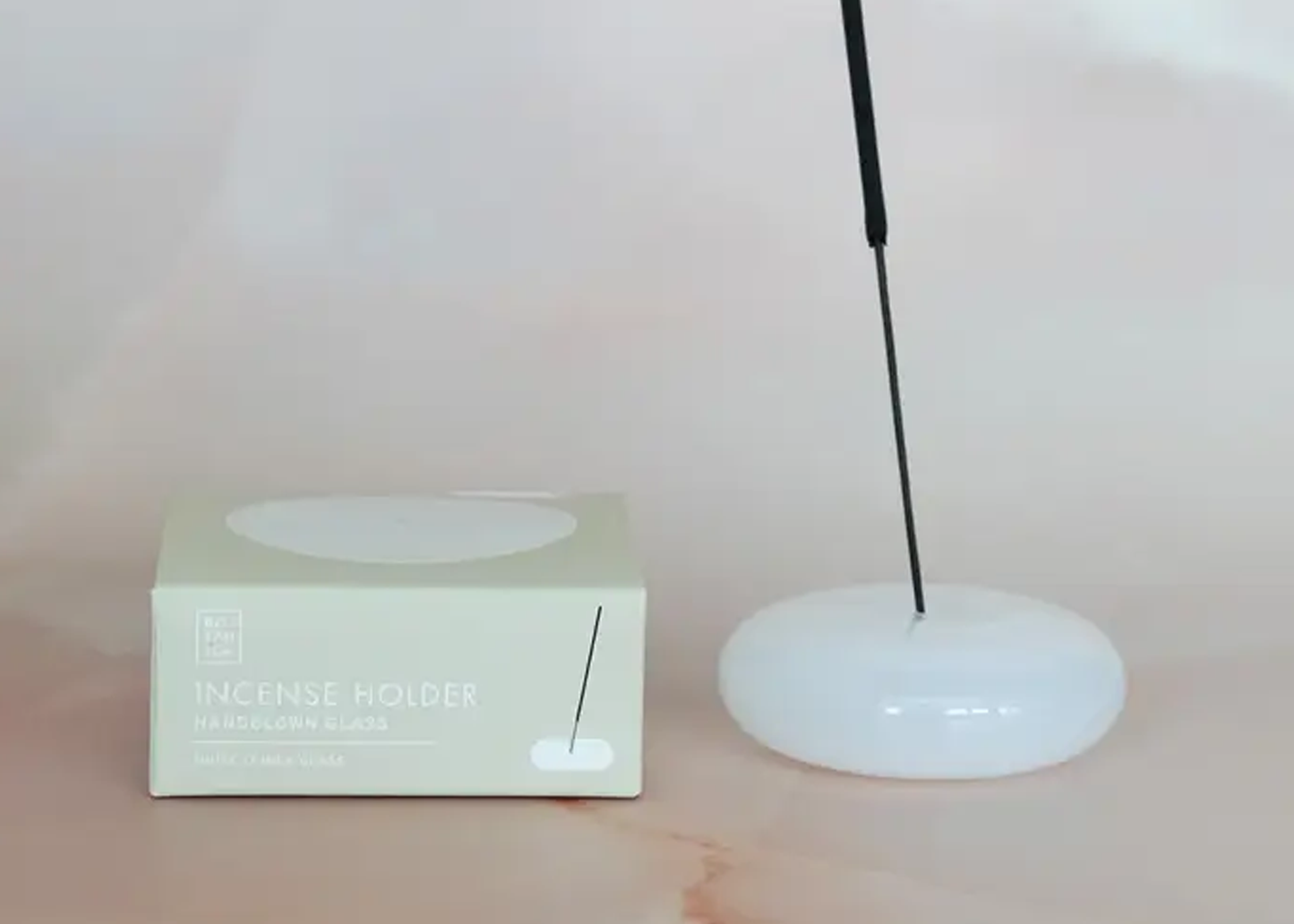 
                  
                    Incense holder with a black incense stick next to its packaging on a light pink background
                  
                