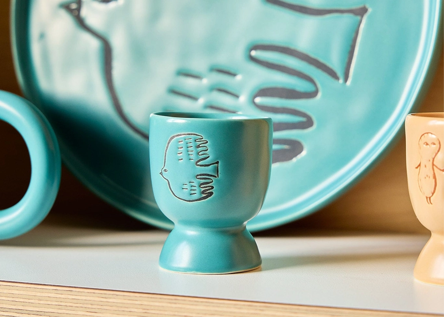 
                  
                    A turquoise ceramic egg cup with an embossed bird design, displayed on a light wooden surface with other matching ceramic dishes in the background.
                  
                