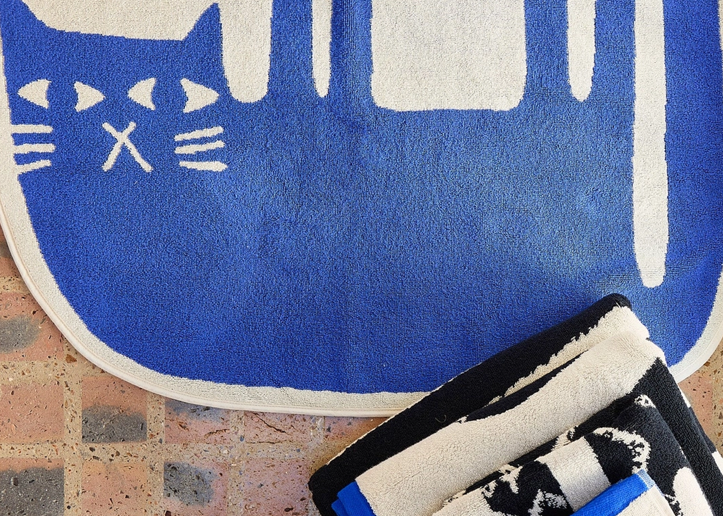 
                  
                    A blue and white cat-shaped bath mat on a tiled floor, with folded towels partially visible in the lower right corner.
                  
                