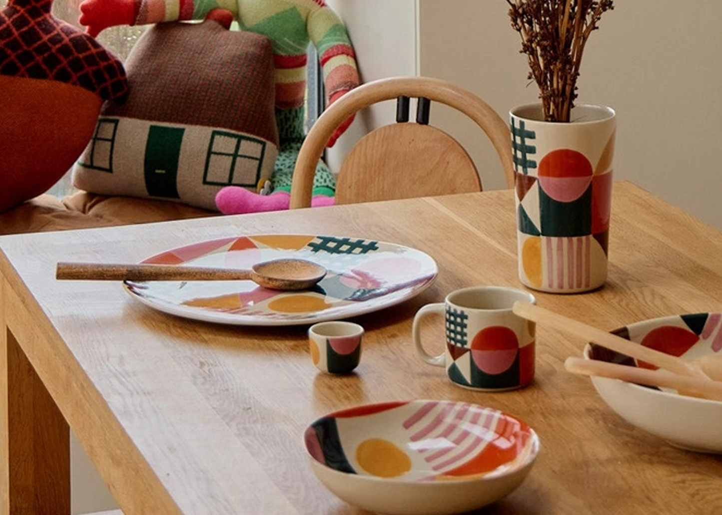 
                  
                    A coordinated ceramic table setting with a plate, mug, small cup, bowl, and vase, all decorated with abstract geometric shapes in orange, red, green, black, and cream, arranged on a light wood dining table in a styled interior.
                  
                
