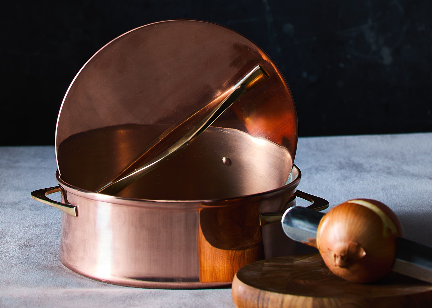 
                  
                    A polished copper stockpot with its lid propped open, resting on a gray surface next to a whole onion on a wooden cutting board. The background is a dark, textured studio backdrop.
                  
                