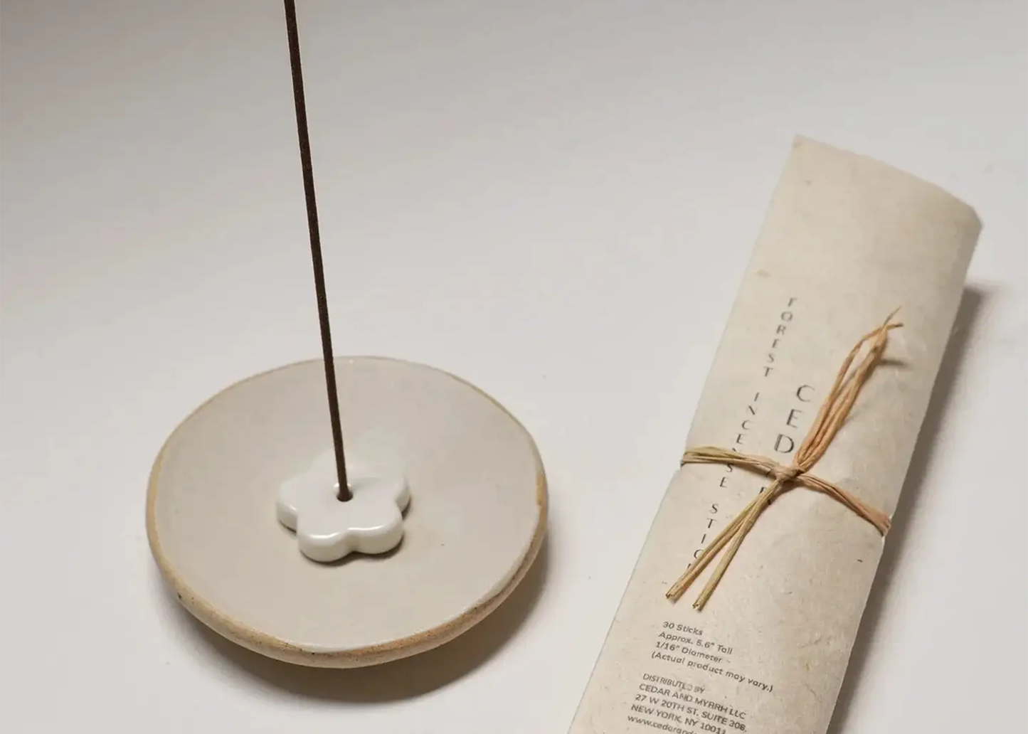 
                  
                    Ceramic incense holder with a single stick of incense on a white surface next to a rolled-up paper with text.
                  
                