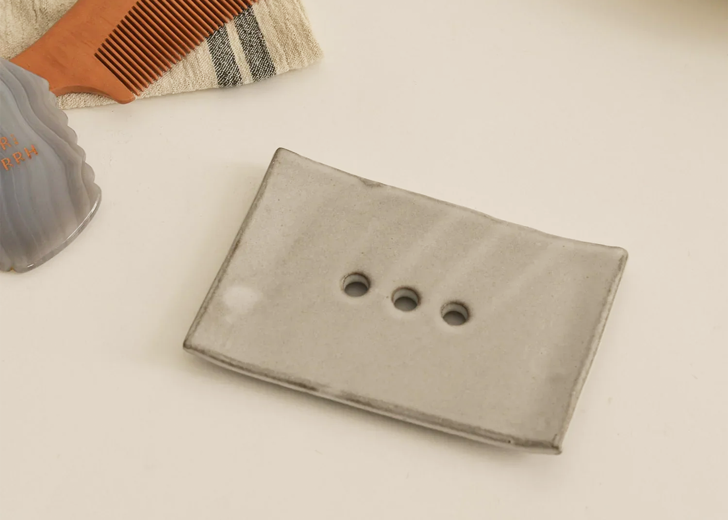 
                  
                    Gray soap holder with three circular holes on a beige surface, with a comb and gray container in the background.
                  
                