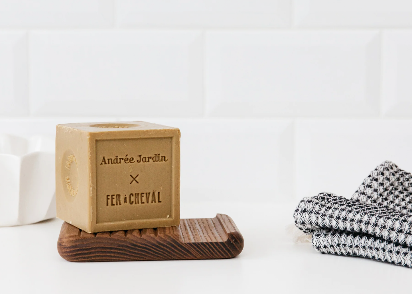 
                  
                    Bar of soap on a wooden stand with 'Andrée Jardin' branding, next to folded towels on a white background.
                  
                