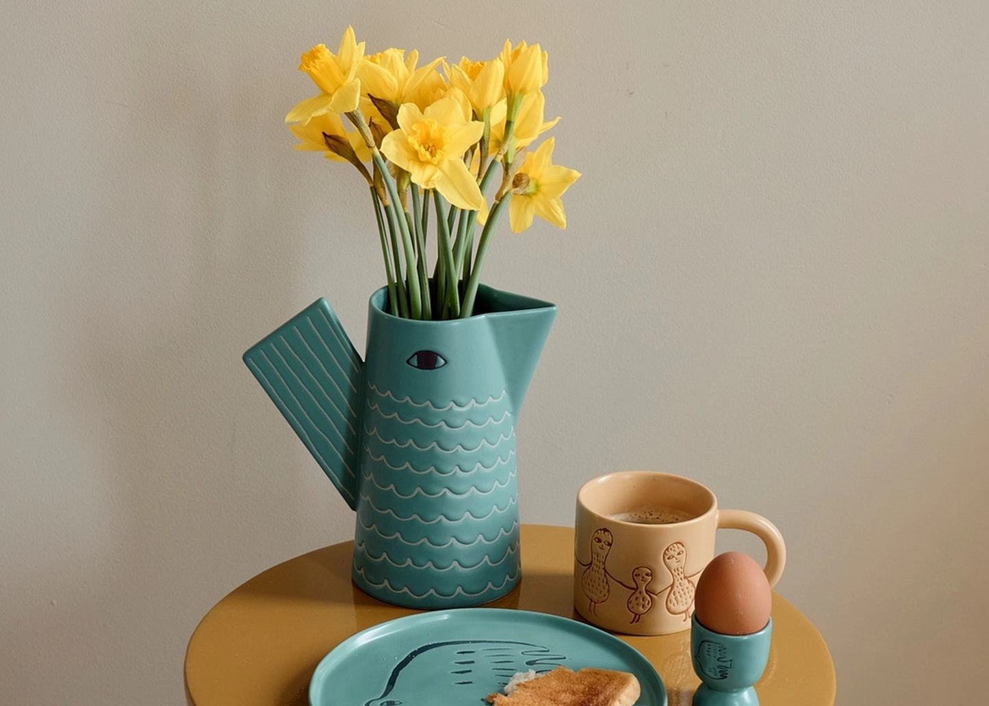 
                  
                    A teal ceramic pitcher vase filled with yellow daffodils, styled on a round mustard-yellow side table alongside a beige ceramic mug, an egg in an egg cup, and a plate with toast against a plain beige wall.
                  
                