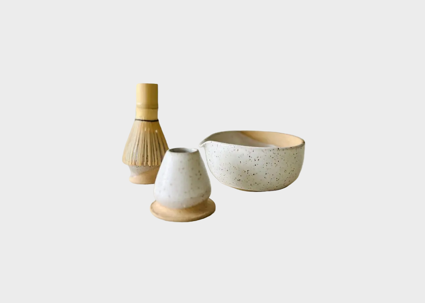 
                  
                    Three-piece ceramic tea set with a speckled cream glaze and natural wood accents, including a bowl, a small vase, and a whisk, on a light gray background.
                  
                