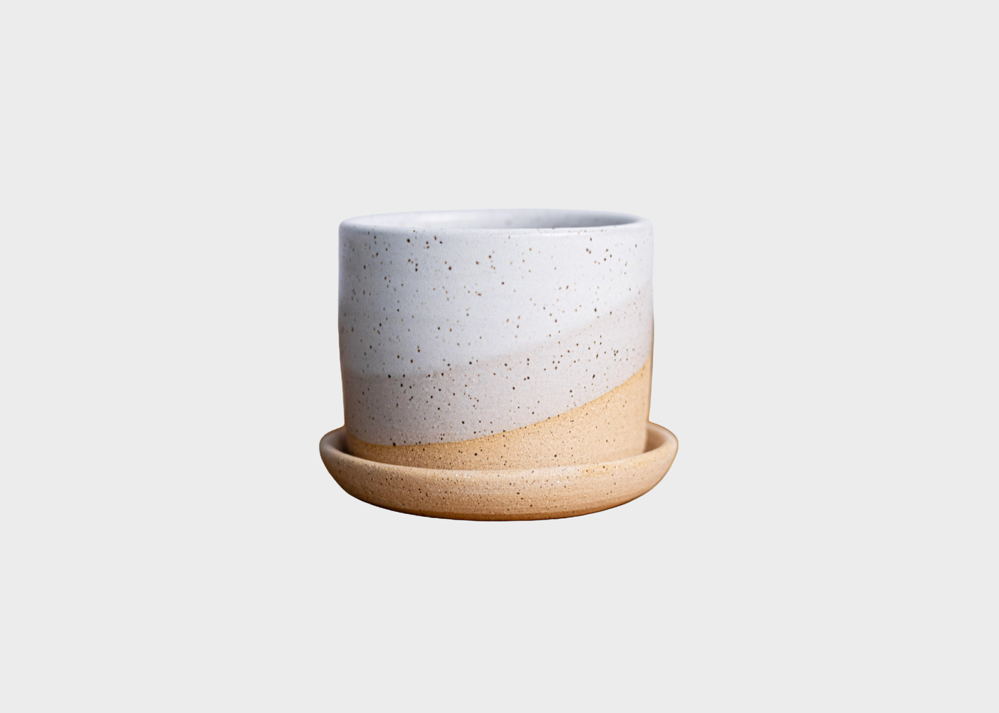 
                  
                    A ceramic planter pot with a matching saucer, featuring a two-tone design with a speckled white upper half and a warm beige lower half, set against a solid light gray background.
                  
                