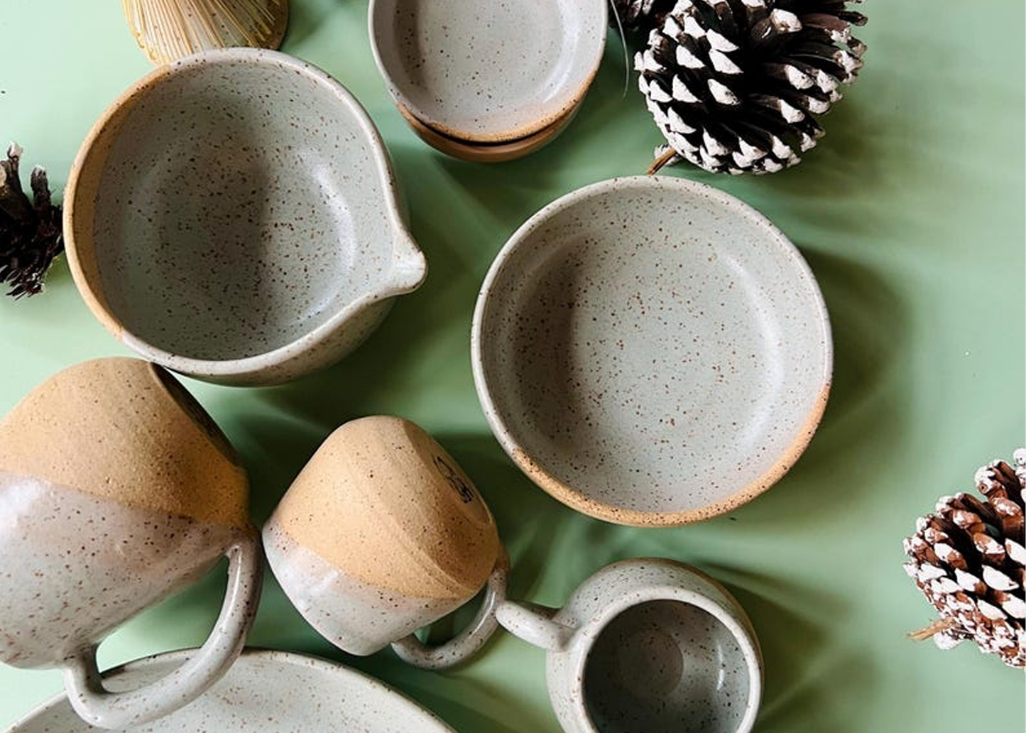 
                  
                    A set of handmade ceramic tableware, including bowls, mugs, and a pitcher, arranged in a flat-lay on a light green background with pinecones.
                  
                