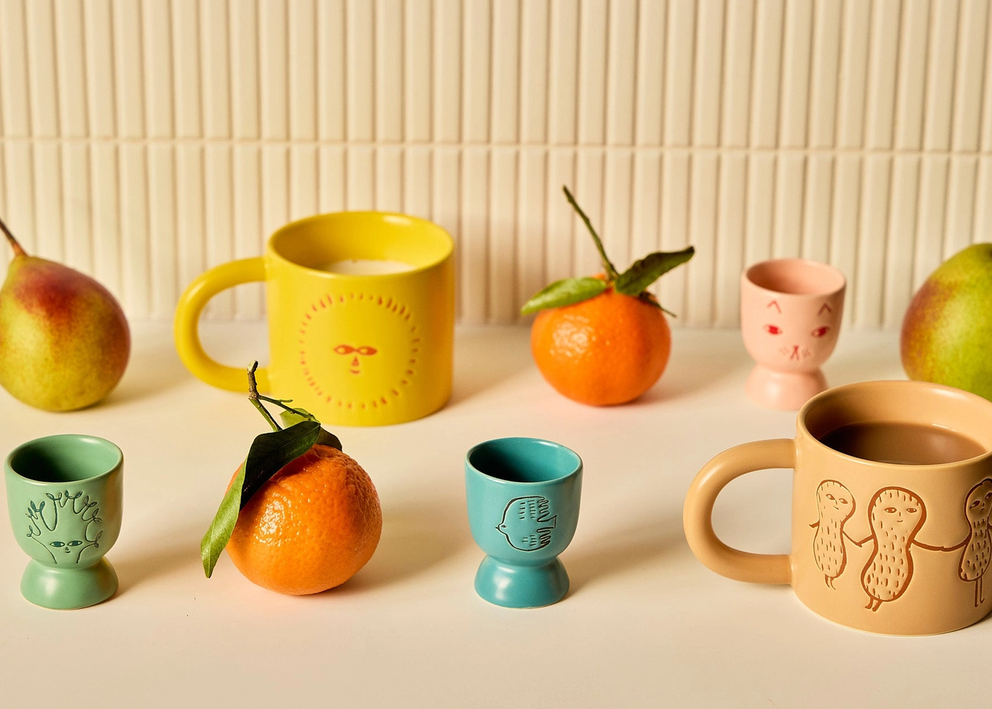 
                  
                    A styled tabletop arrangement of colorful ceramic mugs and small cups with fruit props, including pears and oranges, against a cream ribbed wall background.
                  
                