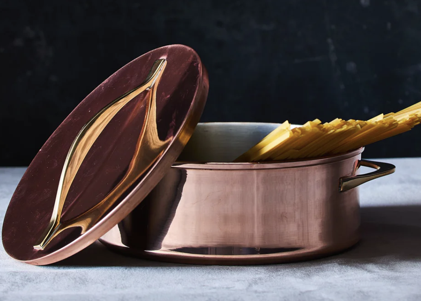 
                  
                    A copper-colored metal pot with a matching lid featuring a glossy, wavy gold pattern, shown with dried pasta inside on a dark textured background.
                  
                