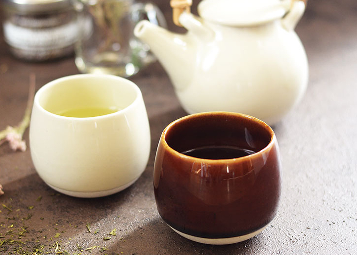 
                  
                    Two ceramic cups, one white and one brown, with a teapot in the background on a textured surface.
                  
                