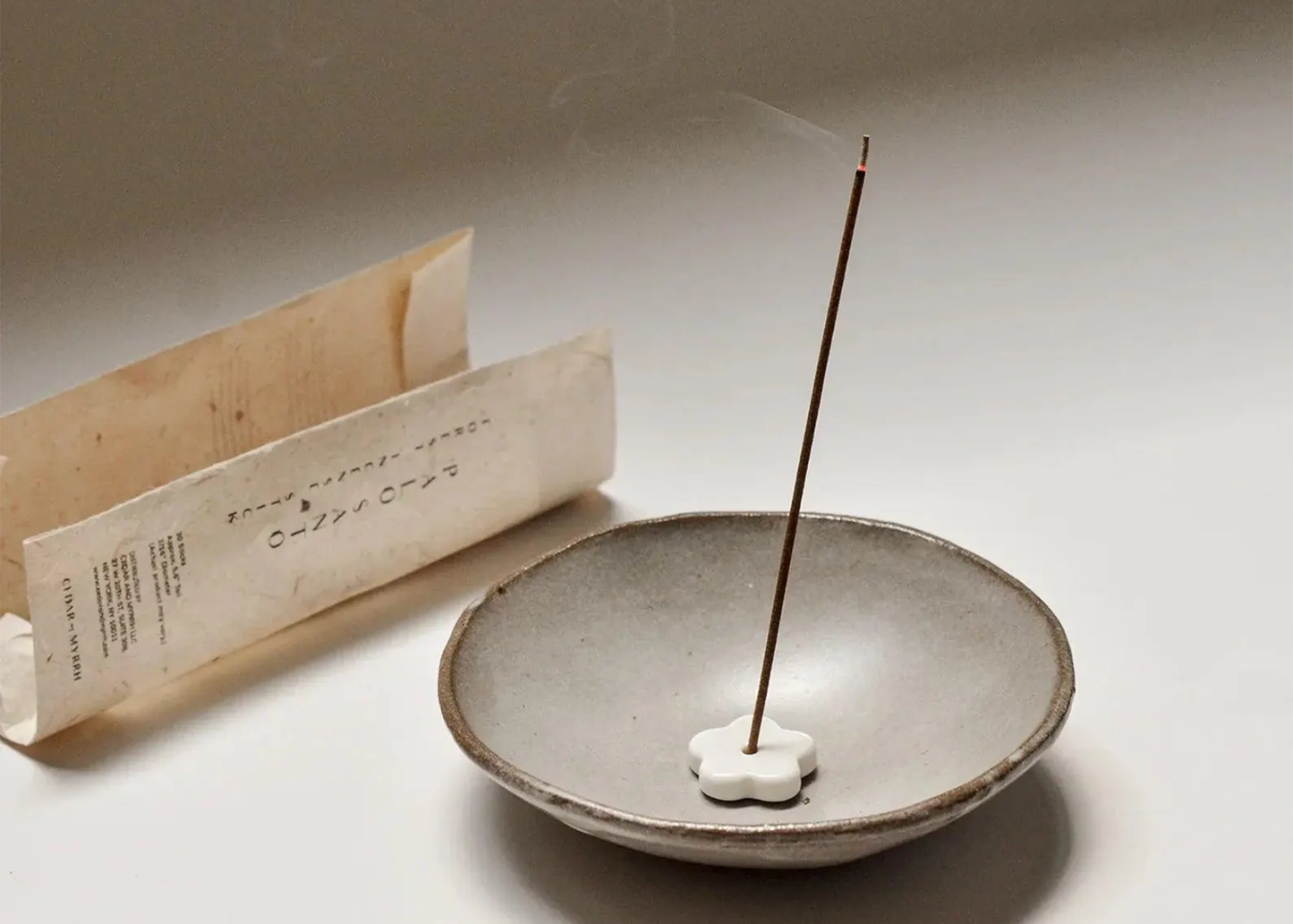 
                  
                    Incense stick burning in a ceramic dish with a paper packet of incense sticks on a light background
                  
                