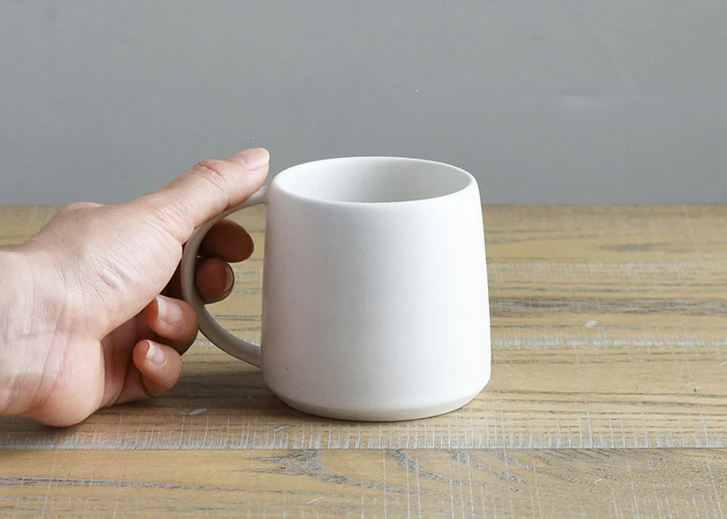 
                  
                    Hand holding a white ceramic mug on a wooden surface with a gray background
                  
                