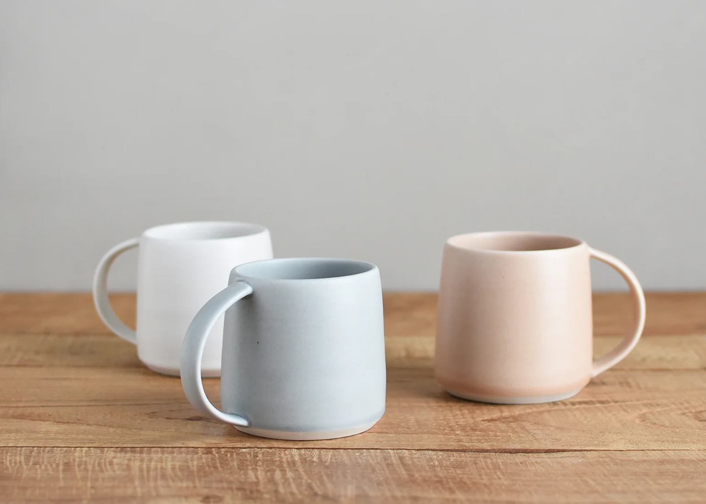 
                  
                    Three ceramic mugs in white, blue, and pink on a wooden surface with a neutral background
                  
                