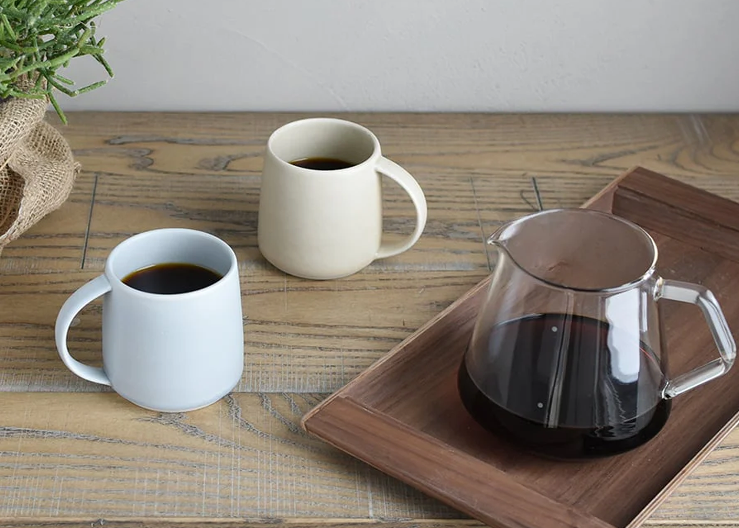 
                  
                    Two mugs and a glass carafe on a wooden surface with a plant in the background.
                  
                