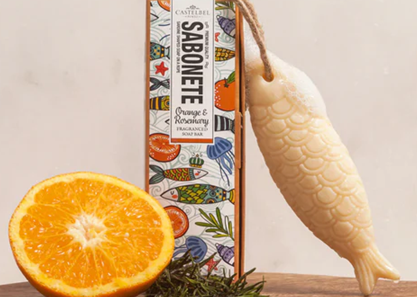 
                  
                    Bar of soap with decorative packaging next to an orange and rosemary on a wooden surface.
                  
                